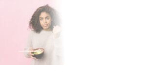 Young woman with curly hair, wearing a light sweater, holding a bowl of Asian cuisine with chopsticks in her right hand, and gesturing with her left hand, standing against a pink background. The right side of the image is blank with a white gradient.