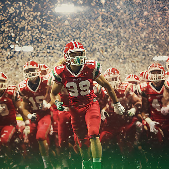 A football team wearing red and white uniforms charges forward on the field under bright stadium lights. The player in the center wears jersey number 88, leading the formation. Confetti fills the air in celebration, and the background shows a large crowd in the stands.