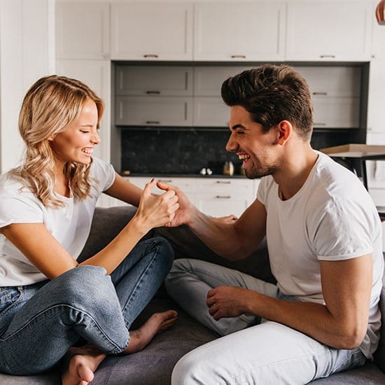 A smiling woman and man sit on a couch in a brightly lit kitchen. They are engaged in a friendly thumb war, both wearing casual white shirts and jeans. The background features white cabinetry and a kitchen island.