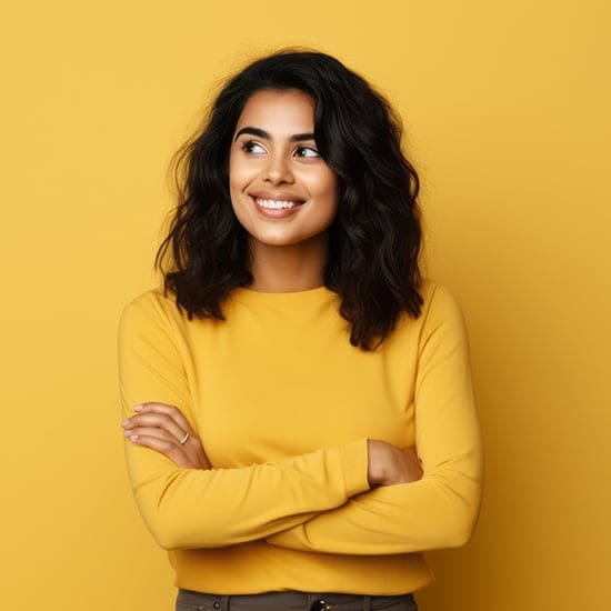 A woman with shoulder-length dark hair smiles while looking to the side. She is wearing a yellow long-sleeved shirt that matches the plain yellow background. Her arms are crossed in a relaxed and confident pose.