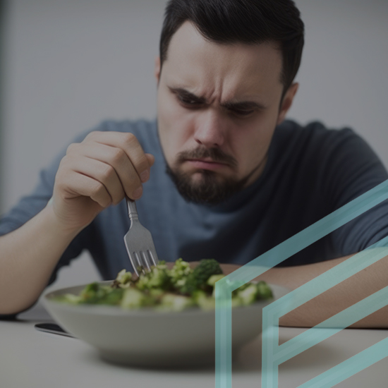 A man with a serious expression sits at a table, holding a fork and looking at a bowl of green vegetables with what appears to be reluctance or displeasure. He is wearing a gray shirt and the background is neutral. A transparent geometric design overlays part of the image.