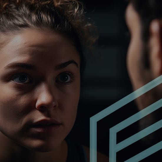 Close-up of a woman with light curly hair looking to the side, her face showing a serious or focused expression. The background is dark and blurred. There is a geometric pattern of light lines overlaying the bottom right corner of the image.