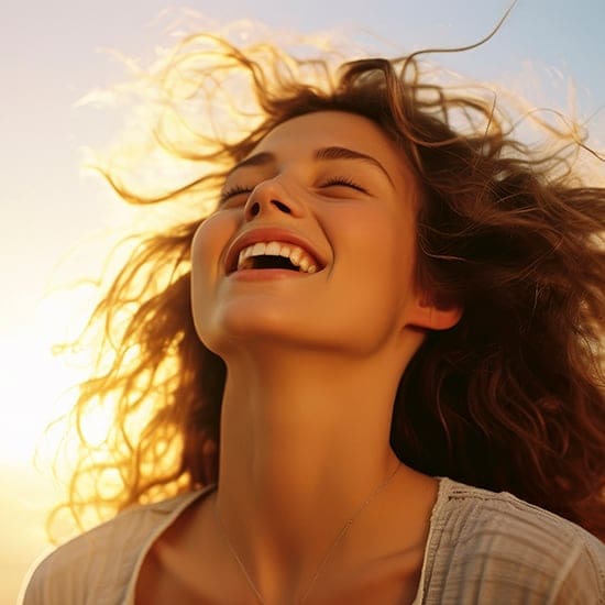 A woman with curly hair joyfully tilts her head back and smiles, basking in the warm sunlight. The golden glow of the sun enhances her radiant expression, and the background is a soft, blurred outdoor setting.