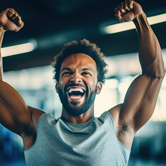 A smiling man with a beard and curly hair, wearing a sleeveless gray shirt, raises his arms in celebration or accomplishment. He is indoors in what appears to be a gym or fitness center, with blurred exercise equipment in the background.