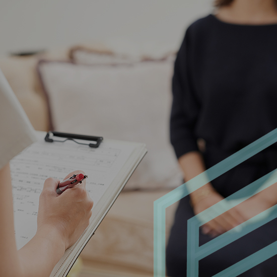 A person takes notes on a clipboard while another individual sits on a couch. The note-taker wears a light-colored top and holds a red pen, while the seated individual is dressed in dark clothing. The background features a blurred view of cushions.