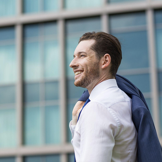 A man is standing outdoors in front of a modern glass building. He is smiling and looking to the side, holding a blue jacket over his shoulder. He is wearing a white dress shirt and a blue tie.