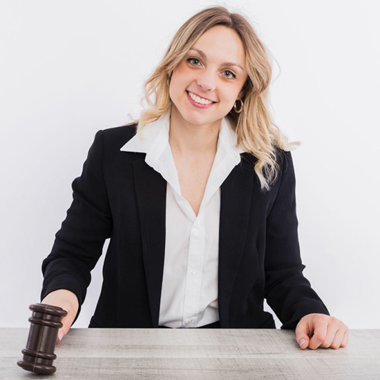 A smiling woman with blonde hair is wearing a black suit and white shirt, sitting at a table with a gavel in her right hand. There is a plain, white background behind her.