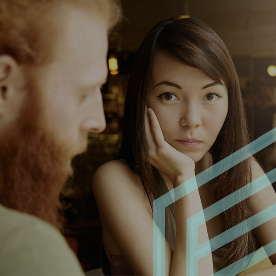 A woman with long brown hair, resting her chin on her hand, looks attentively at a man with a red beard. The man is slightly out of focus. Both are seated indoors, with a geometric pattern partially overlaying the image.