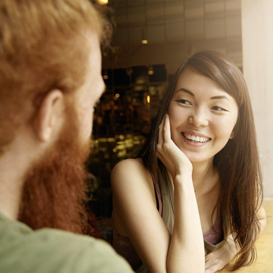 A woman with long hair smiles while resting her chin on her hand, sitting opposite a bearded man in a light-colored shirt. They are in a warmly lit indoor setting, possibly a cafe or restaurant, engaged in a friendly conversation.