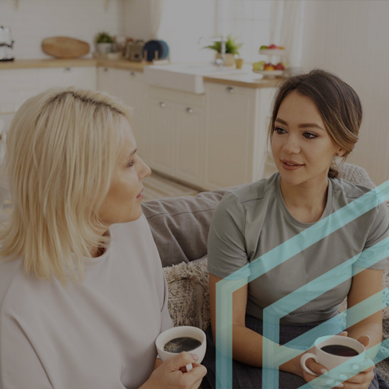 Two women are sitting on a couch in a bright, modern kitchen, engaged in conversation. Both are holding cups of coffee. One woman has blonde hair and the other has brown hair. Light streams in through the windows behind them, enhancing the cozy atmosphere.