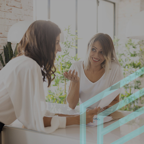 Two women are having a discussion at a desk in a well-lit room with large windows and plants in the background. One woman is sitting while the other stands, gesturing with one hand while holding papers in the other hand. Both appear engaged in the conversation.