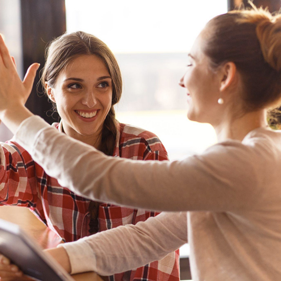 Two women are sitting in a cafe, smiling at each other. One is wearing a red plaid shirt and the other a beige sweater. They appear to be engaged in a lively conversation, with the woman in the plaid shirt raising her hand in a high-five gesture.