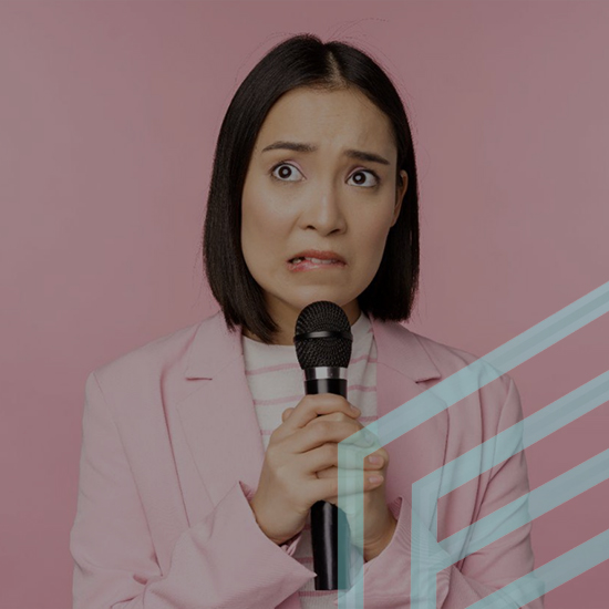 A person with shoulder-length black hair stands against a pink background, holding a microphone. They have a nervous expression, with wide eyes and slightly raised eyebrows. They are wearing a light pink blazer over a white top with pink stripes.