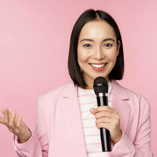 A smiling woman with shoulder-length dark hair is holding a microphone in one hand and gesturing with the other hand. She is wearing a pink blazer over a white and pink striped shirt. The background is solid pink.