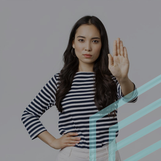 A woman standing and facing the camera, wearing a striped long-sleeve shirt and light-colored pants, raises her right hand with the palm facing outward in a "stop" gesture. She has a serious expression, and there is a turquoise geometric overlay on the right side of the image.