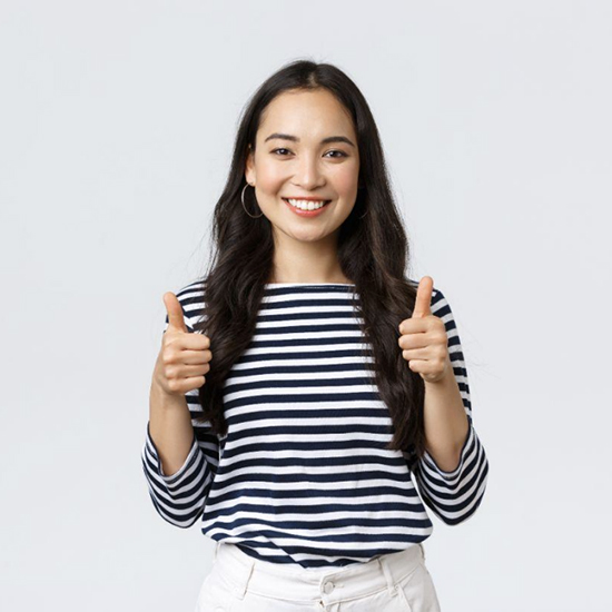 A woman with long dark hair is smiling and giving two thumbs up. She is wearing a white and navy striped shirt and white pants, standing against a plain white background.