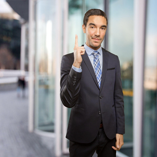 A man in a dark suit with a blue and white striped tie stands outside a modern glass building. He smiles slightly while raising his index finger on his right hand, as if making a point or indicating something. The background is slightly blurred.