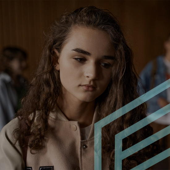 A young girl with long, curly hair looks downward with a sad expression. She is wearing a light-colored jacket with a dark letter 'M' on it. The background is blurry with other people visible, and there is a geometric design overlay on the right side of the image.