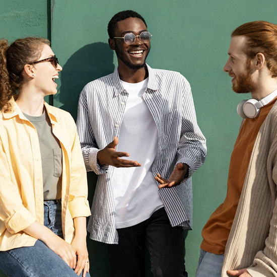 Three friends are standing outdoors against a green wall, smiling and chatting. Two wear sunglasses; one has headphones around his neck. They're dressed casually in light, comfortable clothing, enjoying a moment of laughter in the sunlight.