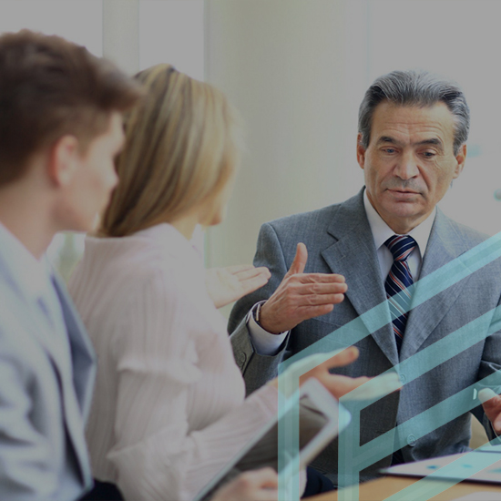 Three individuals are in a business meeting. A man in a gray suit gestures while speaking to a young man and a woman. The woman holds a tablet and is engaged in the conversation, while the young man listens attentively. Light streams in from windows in the background.