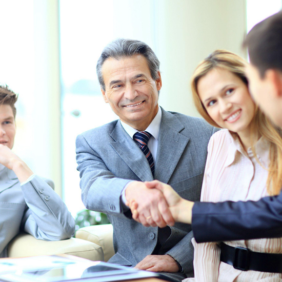 A group of four professionals sitting in a meeting room. The man in the center, wearing a gray suit and striped tie, is smiling and shaking hands with another person. A woman beside him is smiling and looking in the same direction, while another person looks attentively.