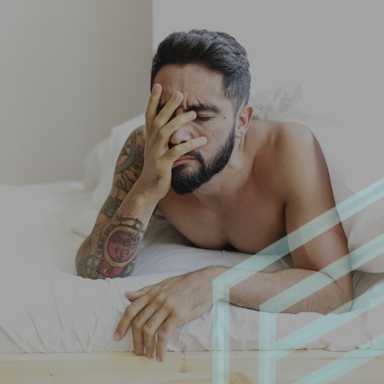 A shirtless man with tattoos is sitting in bed with white sheets, holding his face with one hand and appearing distressed or tired. The background is plain and bright.