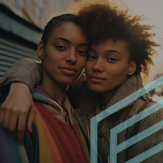 Two people stand closely together outdoors, with one person's arm draped over the other's shoulder. Both are wearing jackets, with one jacket featuring colorful stripes. The lighting suggests a setting sun, casting a warm glow on their faces.