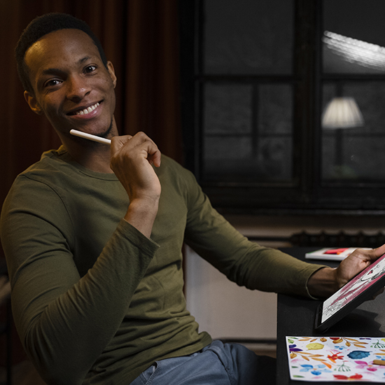 A person wearing a green long-sleeve shirt sits at a table, holding a digital pen and smiling at the camera. They are working on a tablet with colorful designs visible on the surface. The room has dim lighting with a window and curtains in the background.