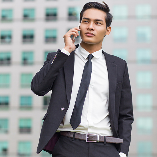 A man dressed in a black suit and tie is standing outdoors in front of a modern building facade, using a smartphone. He looks to the side with a slight expression of focus and contemplation.