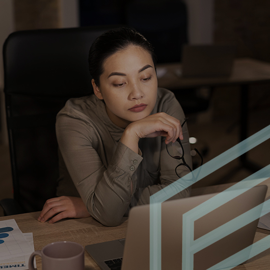 A woman with long dark hair, holding her glasses, sits at a desk looking thoughtfully at her laptop screen. There are papers and a coffee mug on the desk in front of her. The background shows a dimly lit office space with another laptop in the distance.