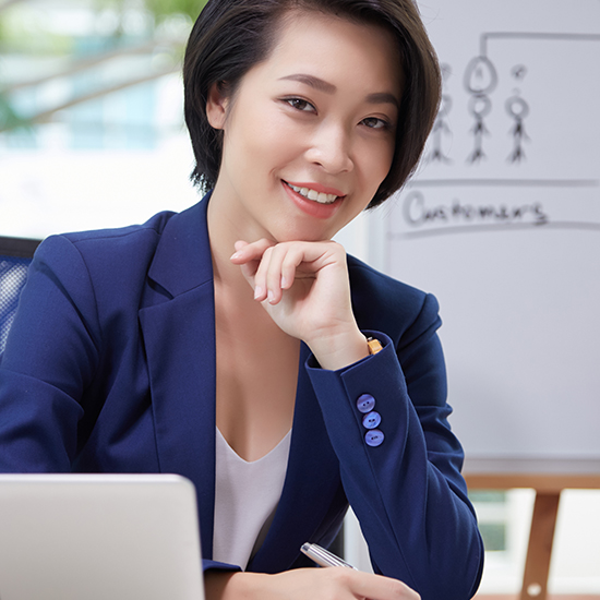 A woman with short black hair, wearing a blue blazer and white top, smiles while seated at a desk. Behind her, there is a whiteboard with a simple diagram and the word "Customers." Her hand rests on her chin, and in front of her is a laptop.