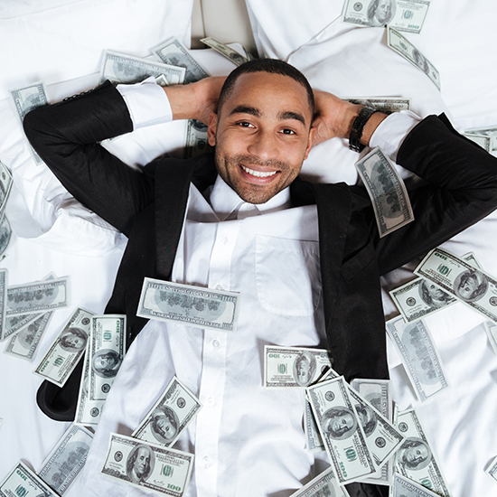 A man lying on a bed with hands behind his head, smiling, wearing a black suit jacket and white shirt. Multiple $100 bills are scattered around and on him.