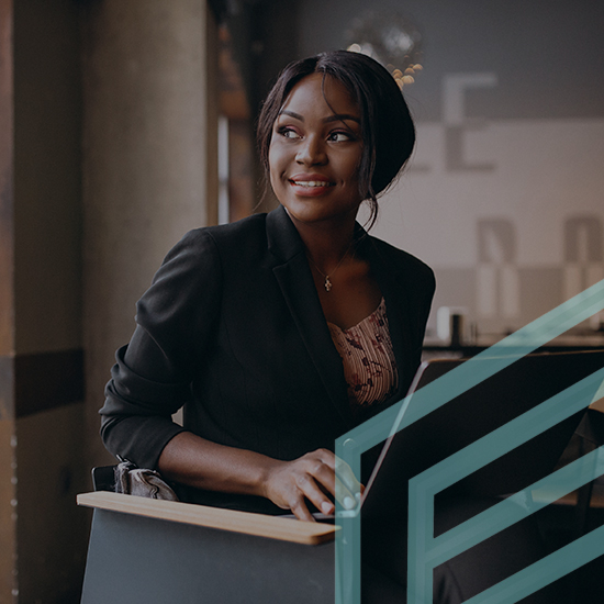 A smiling woman wearing a black blazer and a patterned top sits at a desk, holding a laptop. She looks to her left, appearing thoughtful. The setting is a modern, well-lit indoor space with a muted background.