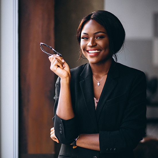 A woman wearing a black blazer and holding a pair of glasses stands by a window. She has a big smile on her face and her arms are crossed. She appears confident and professional, with straight, dark hair tied back.