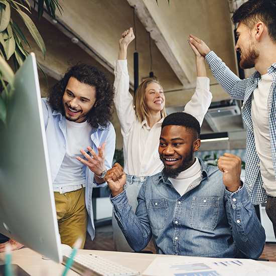 A group of four diverse colleagues celebrate in an office. One man sits at a computer with clenched fists in triumph, while another stands and cheers beside him. A woman and another man in the background raise their hands in excitement, smiling and high-fiving.