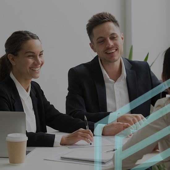 Two professionals, a man and a woman, are engaged in a discussion with a third person across a table. They are seated in an office setting with laptops and a coffee cup on the table, smiling and appearing to be in a positive conversation.