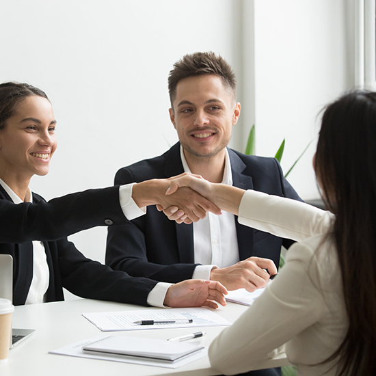 Three people in business attire are engaged in a meeting in an office. A woman on the left and a woman on the right are shaking hands across the table, smiling. A man in the middle is also smiling. The table has papers, notebooks, pens, and a disposable coffee cup.