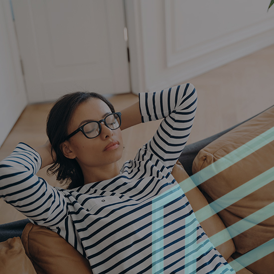 A person wearing glasses and a striped shirt is reclining on a couch with their arms behind their head and eyes closed. The background shows a light-colored wall and a door. The image suggests a peaceful, relaxed moment.