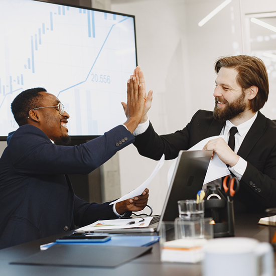 Two businessmen in suits high-five each other in an office. One holds documents, and a laptop is open on the desk. A large screen in the background displays a graph. The atmosphere appears positive and collaborative.