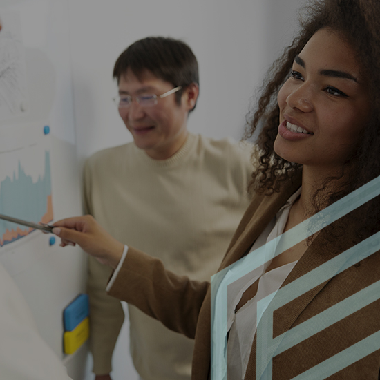 A woman with curly hair in a brown blazer points to a graph on a whiteboard while smiling. A man wearing glasses and a beige sweater stands next to her, also looking at the graph and smiling. The background includes charts and sticky notes on the whiteboard.