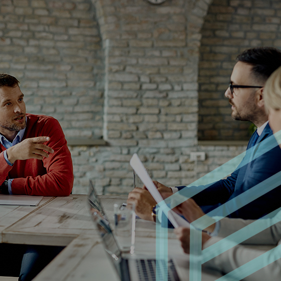 Three people in a meeting room with laptops and documents on the table. The person on the left, wearing a red sweater, is gesturing with their hand. The other two, on the right, are listening attentively. The room has exposed brick walls and a clock is visible in the background.