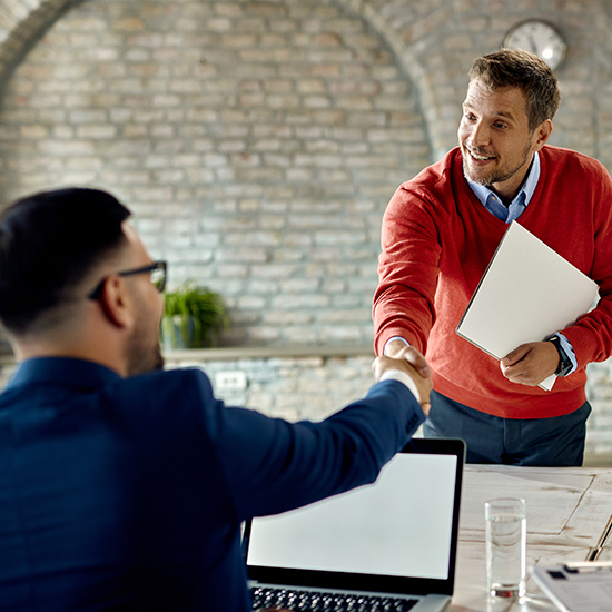 Two men are shaking hands in a modern office. One man, wearing a blue suit, is seated with an open laptop in front of him, while the other, wearing a red sweater and holding documents, is standing. Both are smiling, conveying a sense of agreement or greeting.