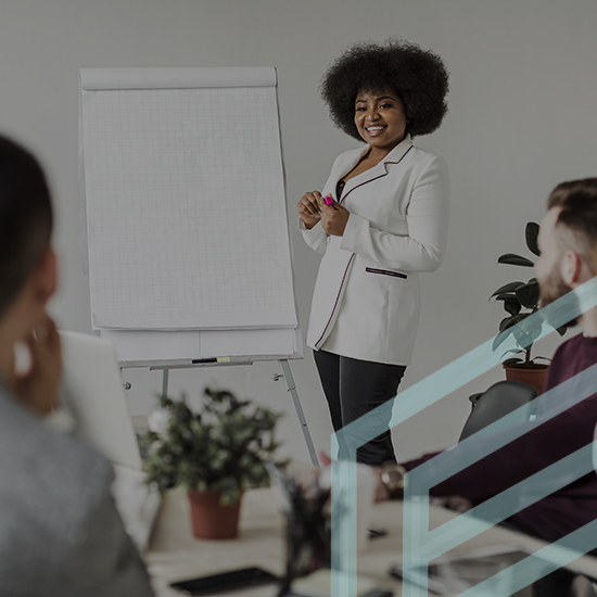 A woman wearing a white blazer stands beside a flip chart, smiling and addressing a group of people seated around a table. The room has plants and office supplies, and the setting appears to be a business meeting or presentation.