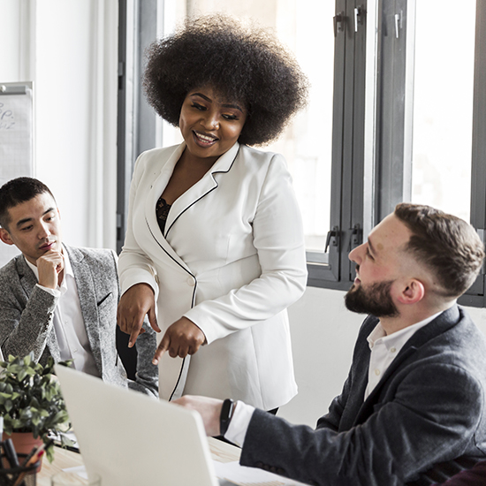 A woman with an afro hairstyle in a white blazer is standing and smiling while pointing at a laptop screen. Two men, dressed in suit jackets, are seated and looking at the laptop. They are in a brightly lit office space with large windows.