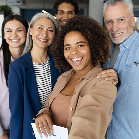 A group of five smiling people is taking a selfie. The group includes a mix of men and women of various ages and ethnicities. They look happy and friendly, with one woman in the foreground holding the camera.