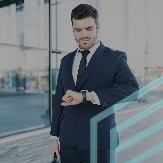 A man in a business suit stands outside a modern glass building, looking at his wristwatch. He holds a briefcase in his other hand, appearing focused and in a hurry. The building's glass reflects the urban environment.