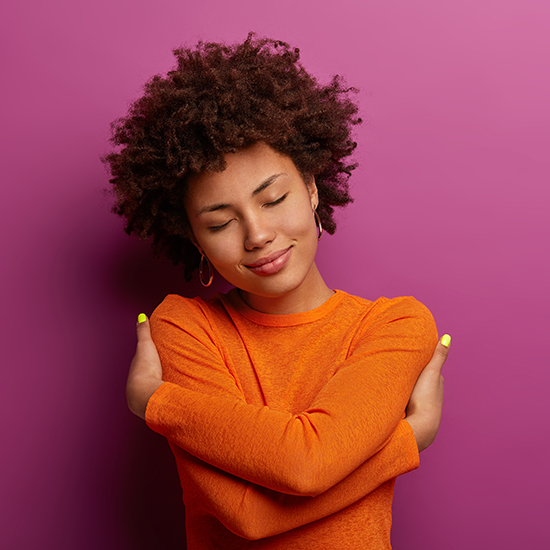 A young person with curly hair embraces themselves, eyes closed, smiling gently. They are wearing a bright orange long-sleeve shirt and small hoop earrings, standing against a plain magenta background.