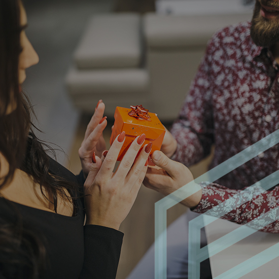 A person in a red patterned shirt presents a small orange gift box adorned with a red bow to another person with long dark hair and red painted nails, wearing a black top. They are indoors, and the image has a partially transparent geometric overlay.