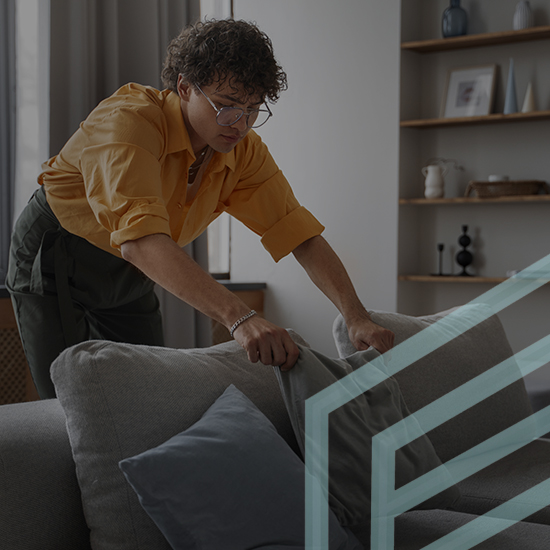 A person wearing a yellow shirt and glasses adjusts the cushions on a gray couch in a modern living room with shelves and decorative items in the background. The scene appears cozy and organized.