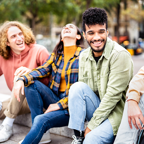 A group of four young adults, three men and one woman, sit together outdoors, smiling and laughing. They appear to be enjoying each other's company. Trees and blurry cityscape are visible in the background, suggesting a casual, urban setting.